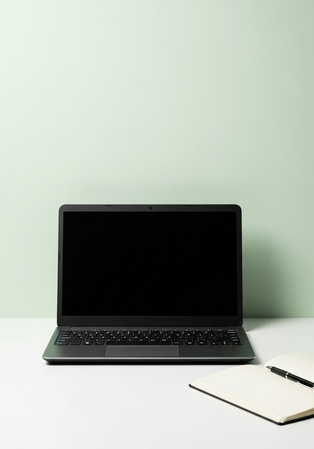 Black laptop, open notebook and pen on a white desk against a light green wall