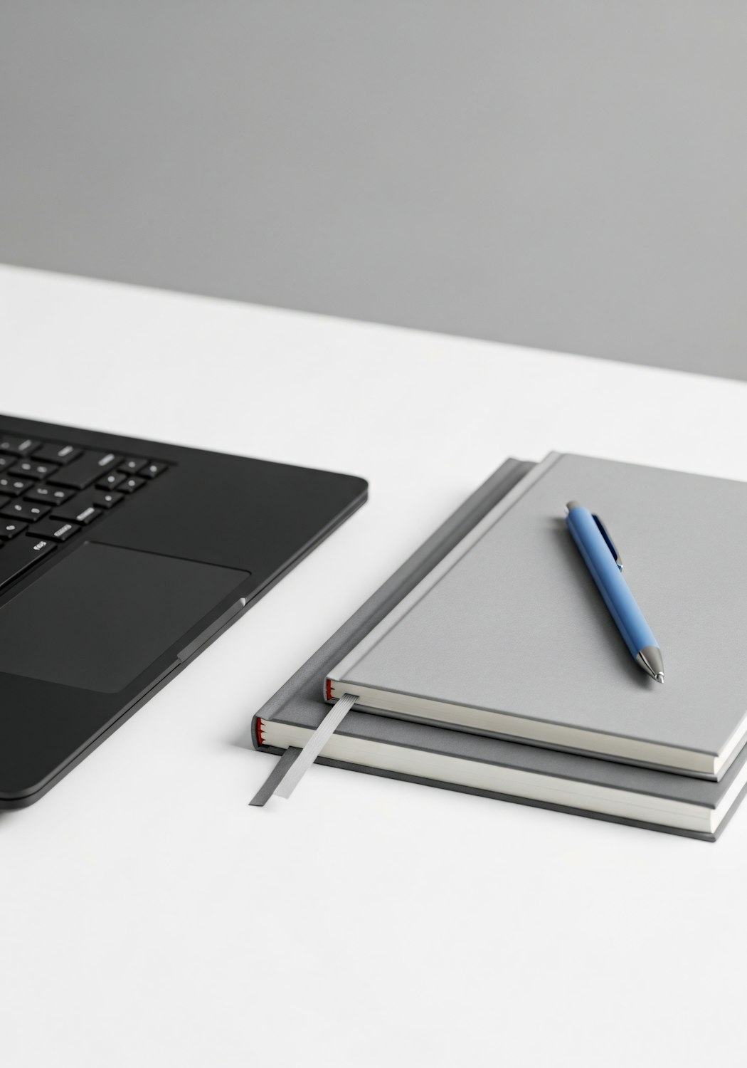 Black laptop, grey notebooks and a blue pen on a white desk
